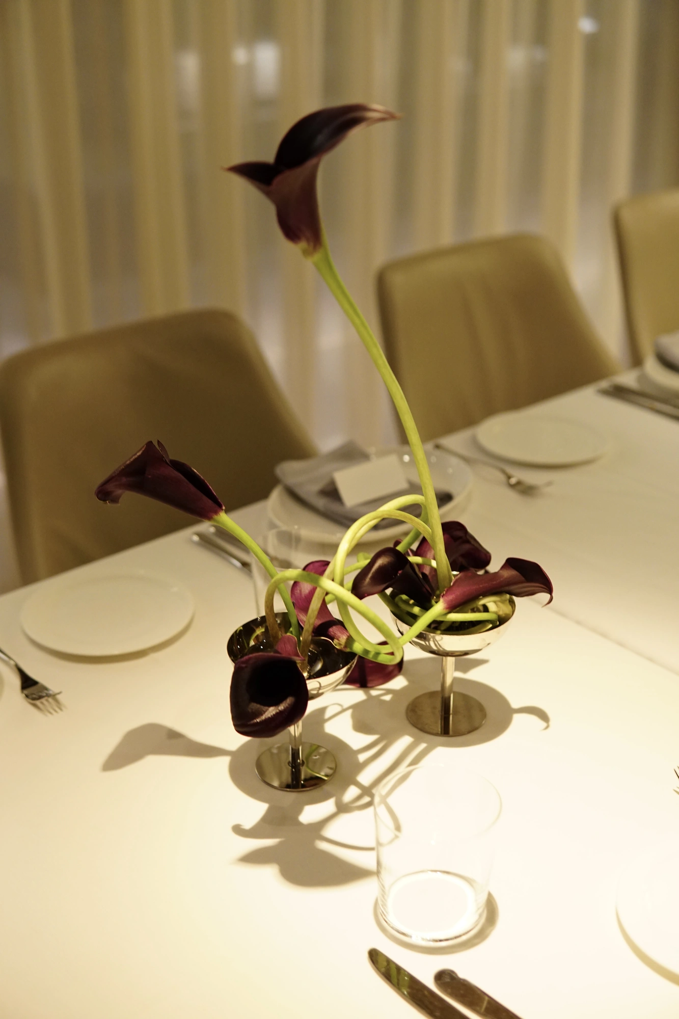 Paired steel cups holding looping dark calla lilies on a set table